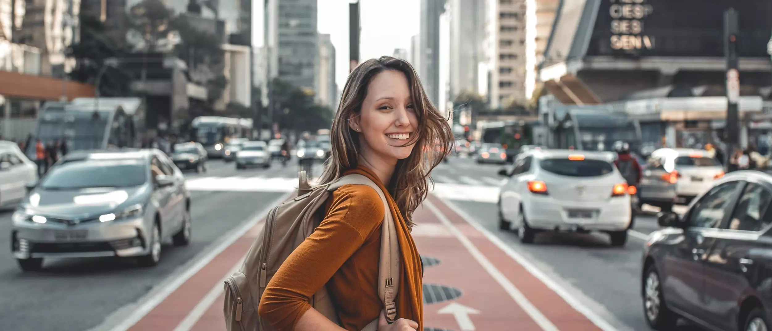 Eine Junge Frau steht auf einer befahrenen Straße in einer Großstadt und grinst in die Kamera
